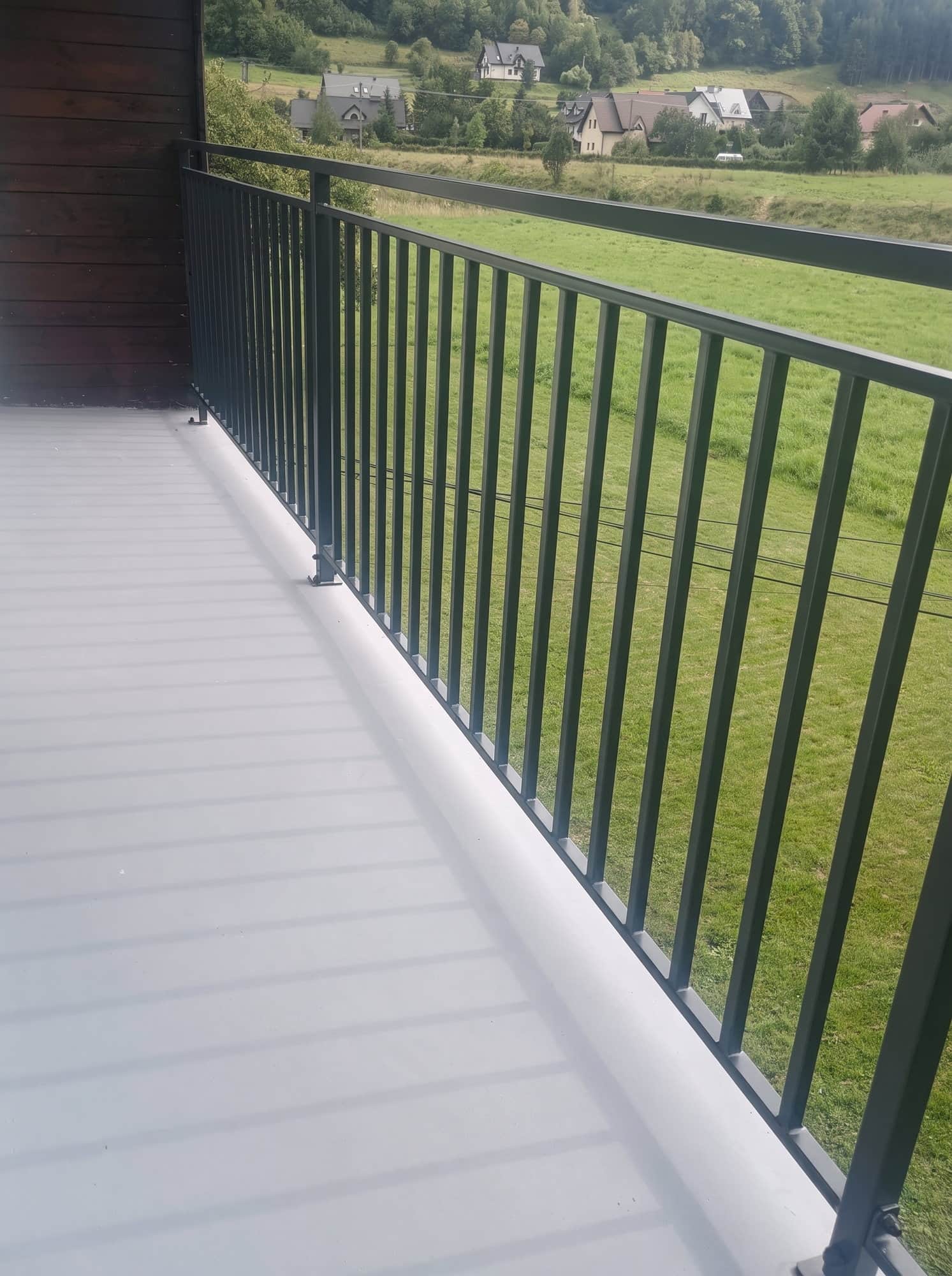 Balustrada balkonowa z pionowych prętów, za nią łąki i wzgórza