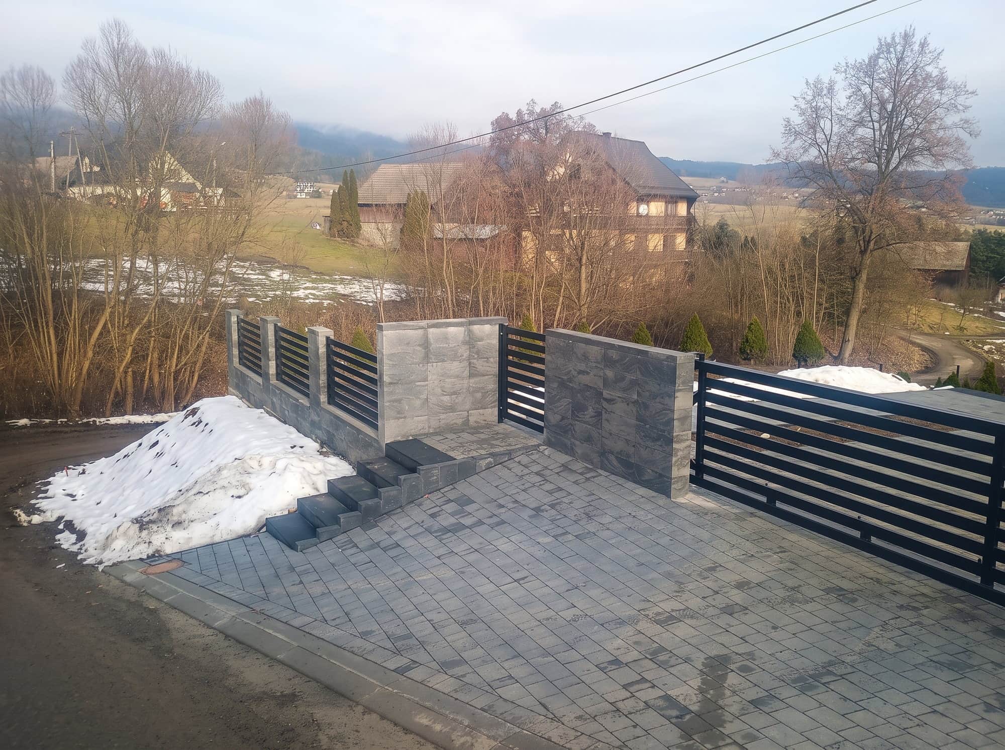 Brama wjazdowa i przęsła z lameli na murze z ciemnego kamienia, zimą, z widokiem na dolinę