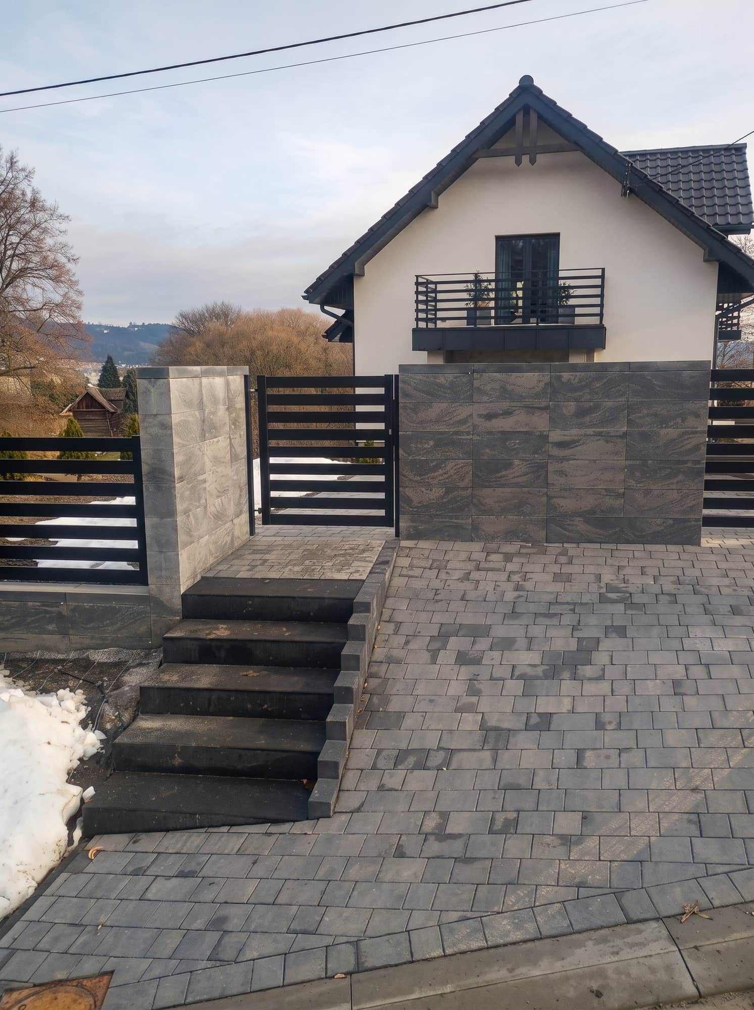 Brama z ciemnych lameli przy słupie obłożonym kamieniem, za nią dom z balkonem