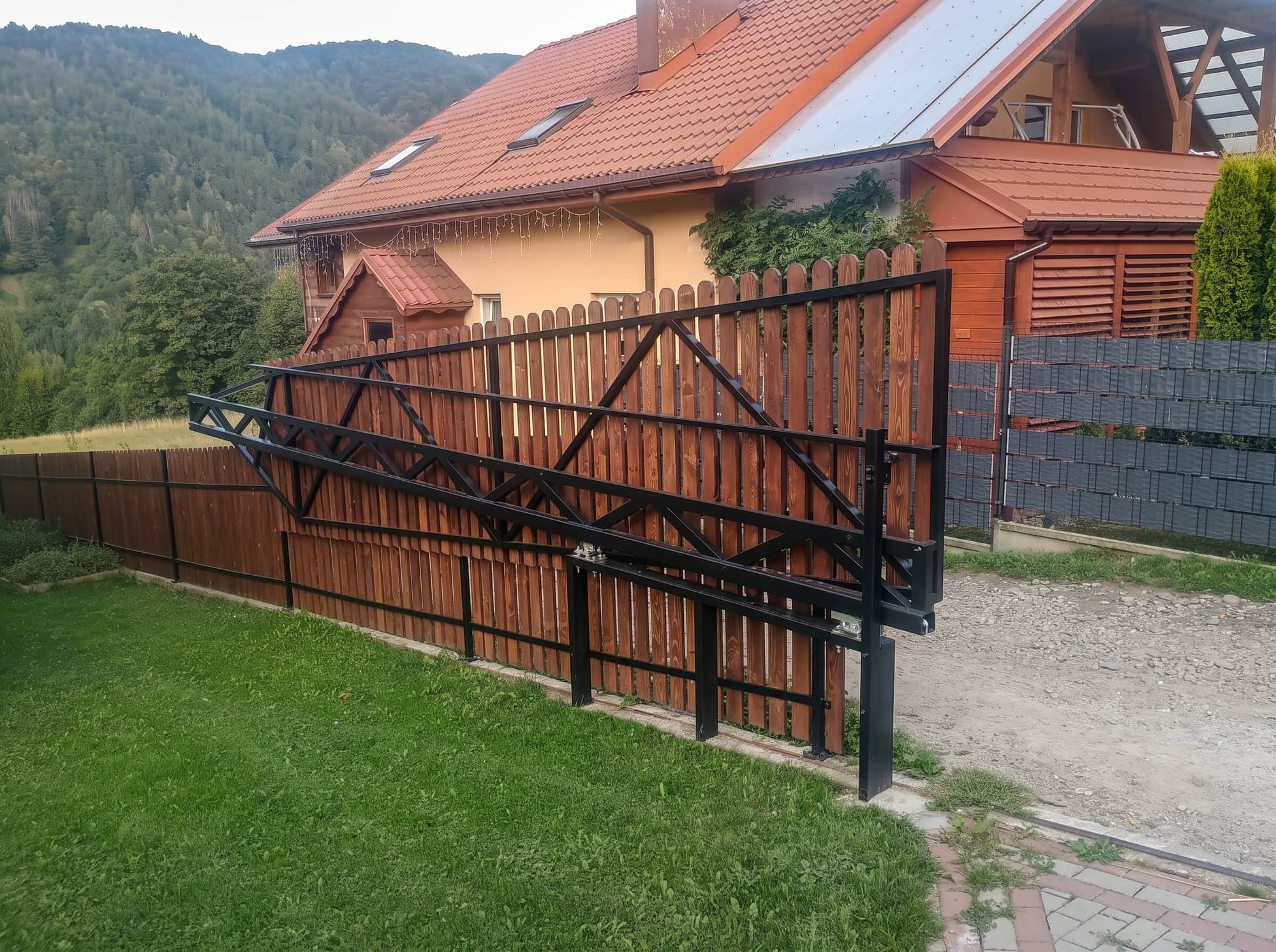 Czarna brama przesuwna o konstrukcji kratownicowej przy drewnianym płocie, w tle zalesione góry